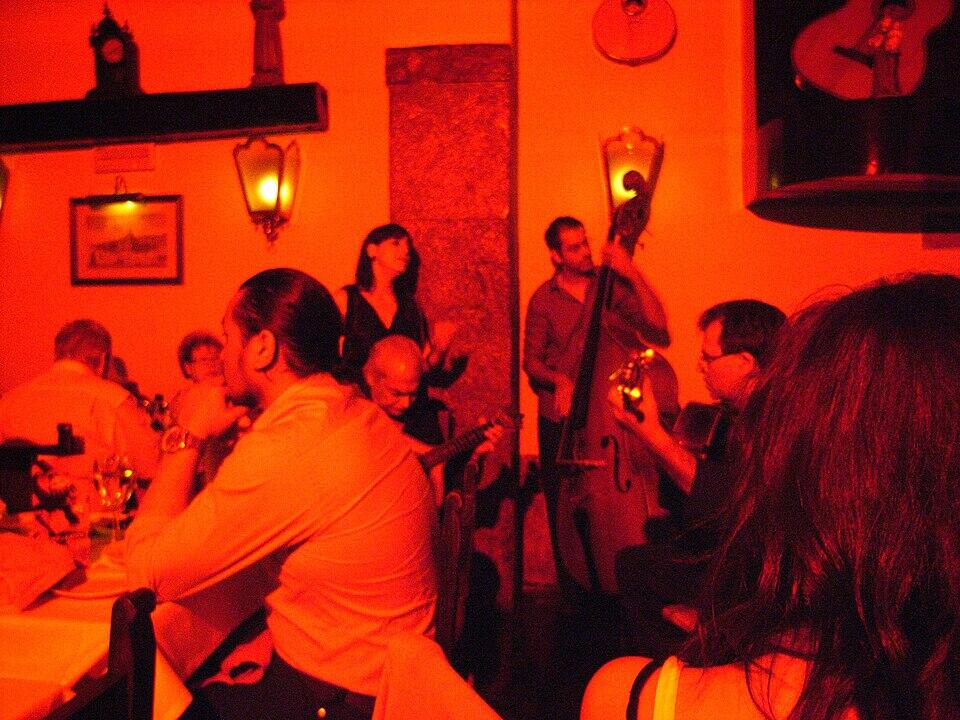 Una escena en un restaurante con poca luz en la que aparecen músicos tocando instrumentos, iluminados con cálidos tonos rojos, y clientes disfrutando del ambiente.
