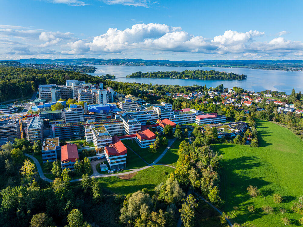Vista a&eacute;rea de un campus universitario amplio y moderno, con edificios de tejados rojos, rodeado de una exuberante vegetaci&oacute;n, junto a un lago tranquilo bajo un cielo azul.
