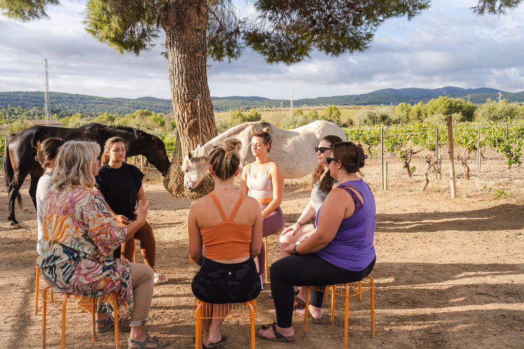 Un grupo de mujeres sentadas en c&iacute;rculo bajo un &aacute;rbol, con caballos pastando cerca en un pintoresco paisaje de vi&ntilde;edos.