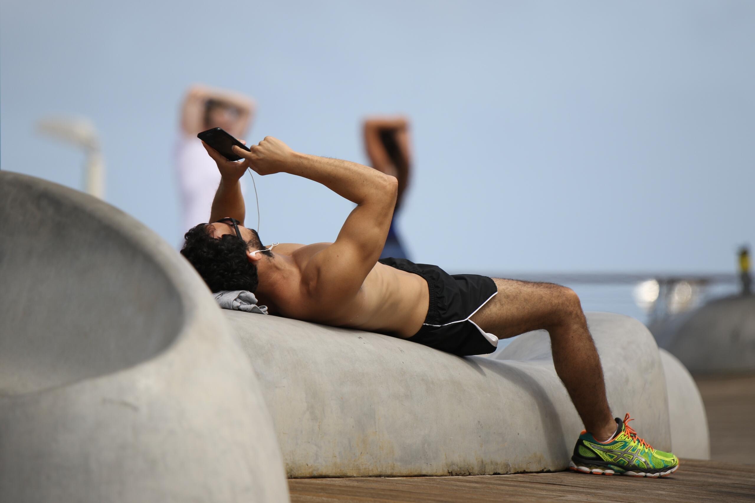 Un hombre tumbado en una cama con un smartphone en la mano, junto al cual hay un reloj inteligente y unas zapatillas de colores.